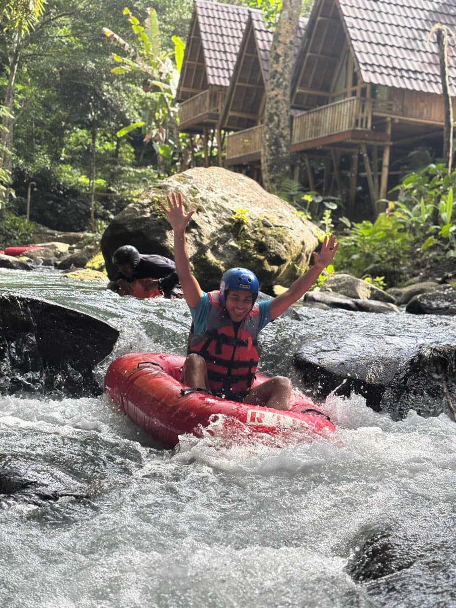 A rider cheers with arms up in front of the riverside bamboo lodges