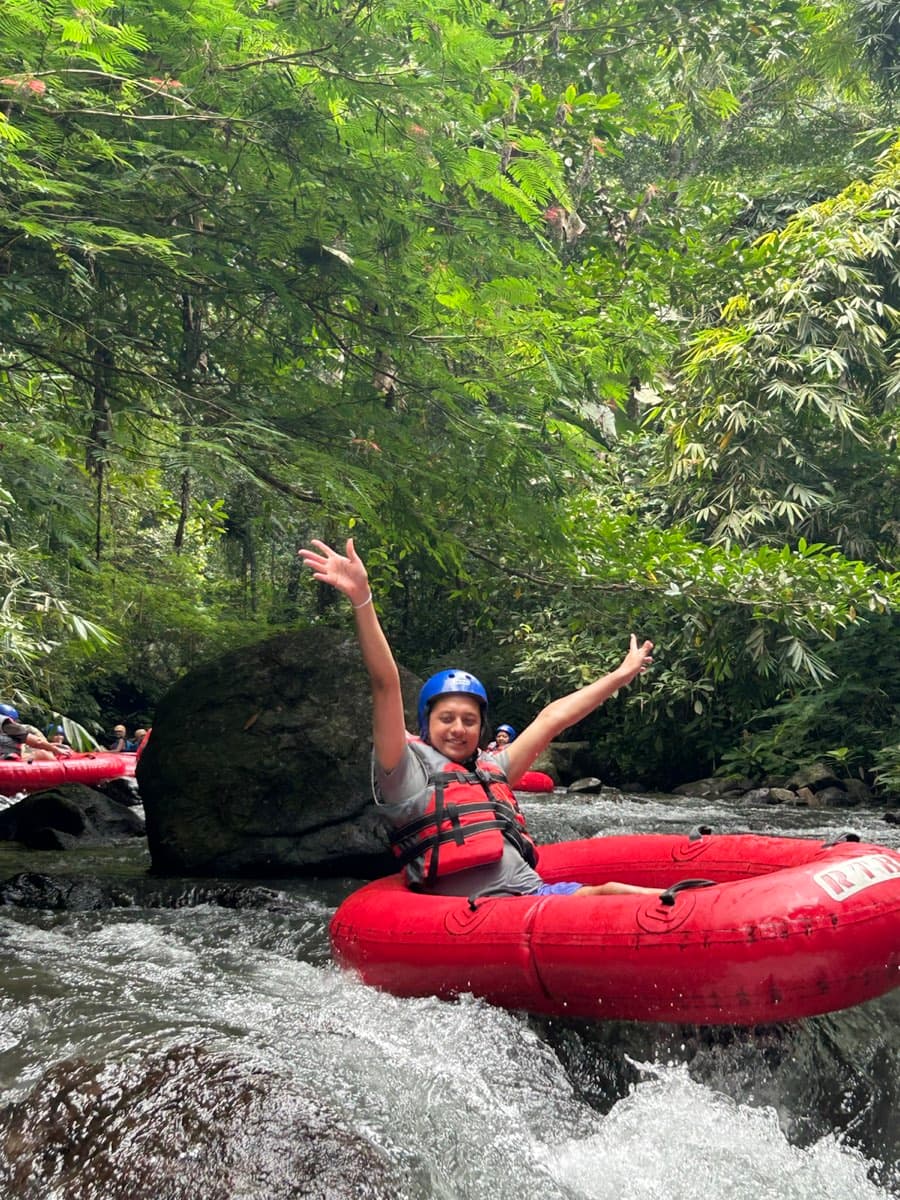 A rider lifts both arms riding a whitewater rapid past a boulder