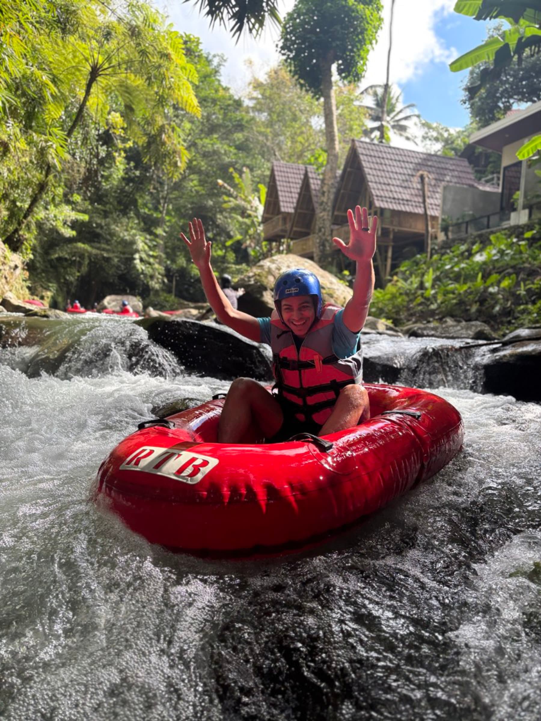 Smiling tuber with arms raised riding a red tube down a sunlit jungle river near Ubud, Bali